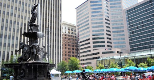 A tall, dark colored fountain on the left side, surrounded by downtown Cincinatti skyscrapers behind it and rows of tables with green umbrellas on the right side.
