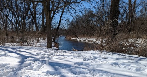 A white, snow covered trail in the foreground flanked by bare trees with a blue river in the background.