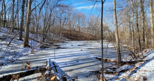 A snow covered frozen river at Highland State Recreation Area surrounded by bare, brown trees under a bright blue sky.