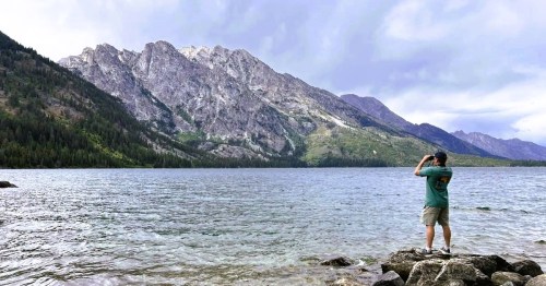 A man taking pics while standing on the rocky edges of an alpine lake in Grand Teton National Park in Wyoming.