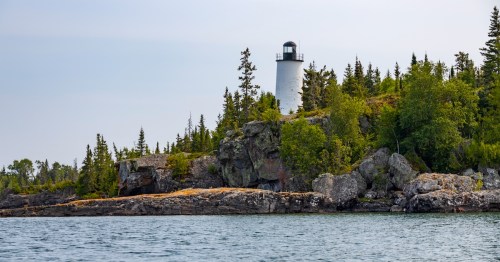 A white lighthouse stands above a rocky shore surrounded by evergreen trees and the blue water of Lake Superior at Isle Royale National Park.