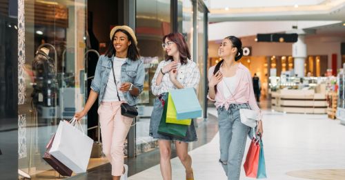 Three young girls are smiling while carrying shopping bags and browsing through a mall.