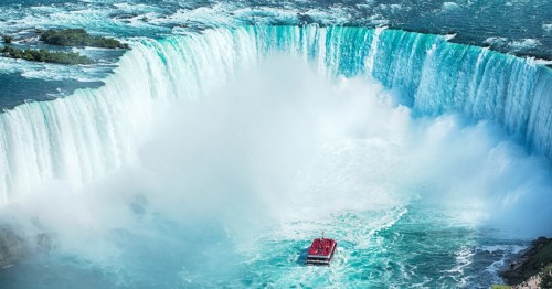 A small red boat floats in front of a wide waterfall and seems dwarfed by the size of the mist coming off of the falls.