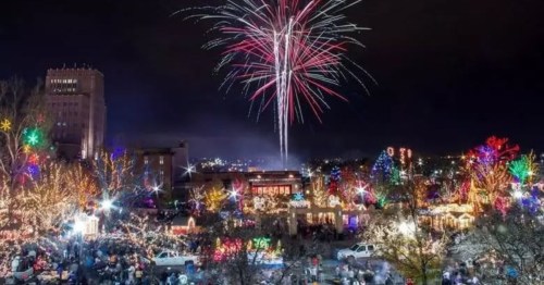 A nighttime view of Ogden's Christmas Village in Utah, featuring vibrant holiday lights, glowing displays, and fireworks above the city skyline.