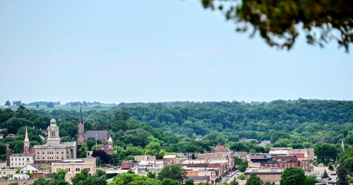 A view of a small, historic town in a green forested river valley beneath a hazy sky.