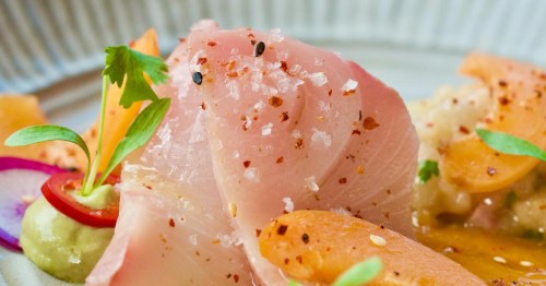 Slices of raw fish, possibly hamachi or tuna, seasoned with coarse salt and chili flakes, garnished with herbs and vegetables. 