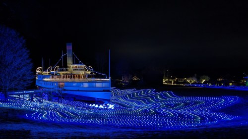 the ground of the shelburne museum is covered in blue lights that look like ocean water and the museum's boat, covered in christmas lights looks like it's floating