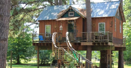 a curved staircase leads up to a cabin on stilts with a wraparound deck in the trees in shreveport Louisiana