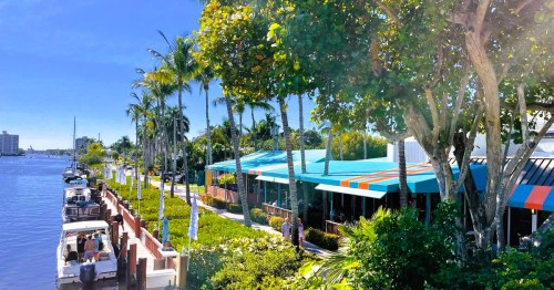 An inlet with several docked boats lined with palm trees and colorful storefronts at a coastal town in Florida.