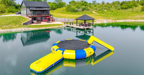 A large, modern black cabin overlooks a pond with a bright yellow and blue inflatable water trampoline and slide.