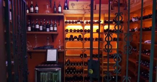 A well-stocked, warmly lit wine cellar or storage room with rows of bottles on wooden shelves and a metal gate. 