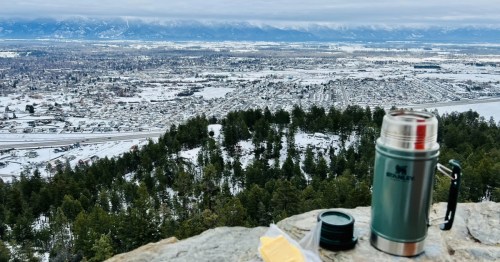 A thermos and baked good sit on a rock railing overlooking the Flathead Valley covered in a blanket of snow.