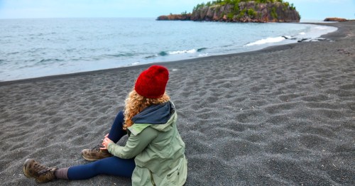 Person wearing a red knit hat and green jacket sitting on a pebble beach, looking out at Lake Superior with a rocky, tree-covered island in the distance.
