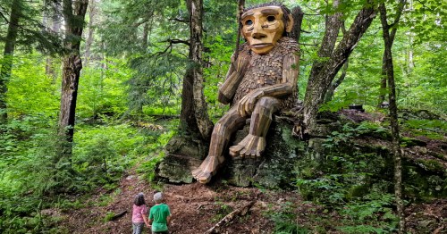 Wooden troll sitting in the forest with people looking up at it.