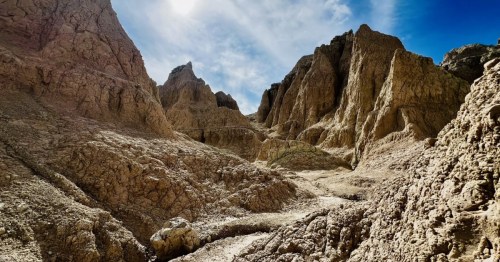 Notch Trail, Badlands National Park. Towering, eroded tan rock formations and a rocky trail under a bright sun and blue sky.
