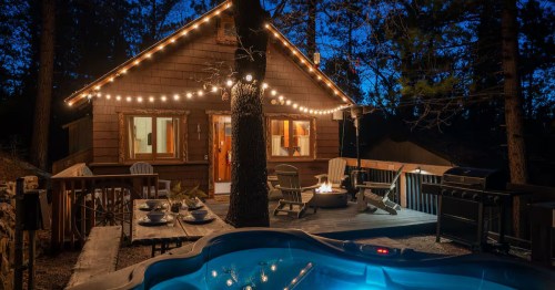 The exterior of a 100-year-old Southern California cabin at night with a hot tub, fire pit, patio seating, and string lights overhead.