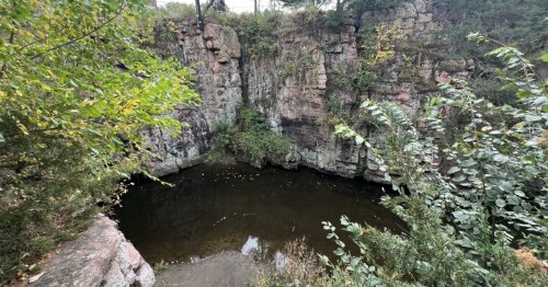 Devil's Gulch, Garretson. A dark pool of water at the base of steep Sioux Quartzite cliffs surrounded by green foliage.