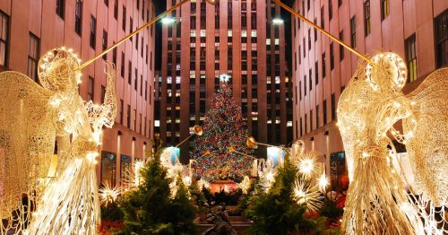An image of Christmas time in New York City in Rockefeller Plaza, where the main entrance is lined with illuminated angels holding trumpets, and there's a beautiful Christmas tree with multicolor lights.