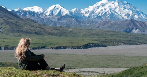 A woman sits in an open Alaska landscape, where large mountains loom in the distance.