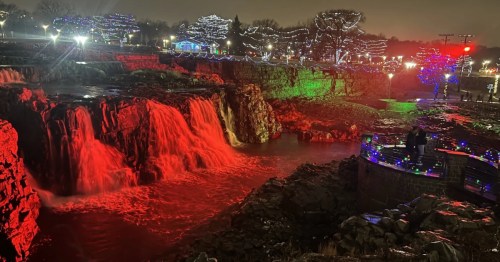 Falls Park, Sioux Falls, illuminated for Christmas. The falls glow bright red and green, with trees wrapped in white lights.