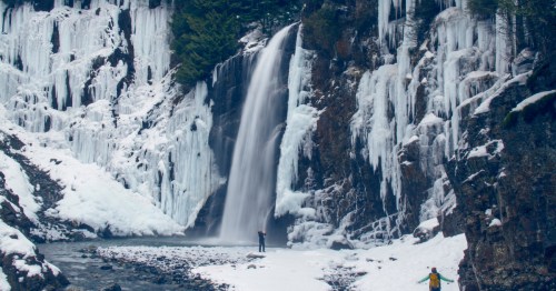 Hikers stand at the base of a large waterfall with massive icicles on the cliff walls.
