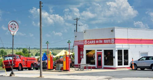 A red truck stands in front of a vintage gas station with a blue sky.