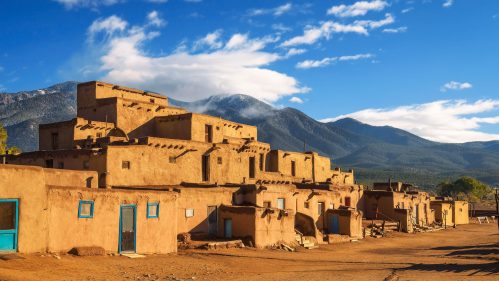 Historic adobe buildings under a blue sky with mountains in the background, showcasing a desert landscape.