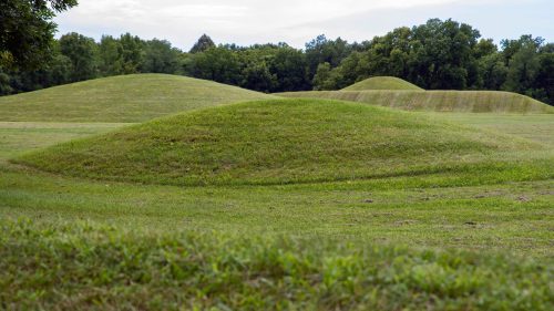 Three grassy mounds rise gently in a green landscape, surrounded by trees under a cloudy sky.