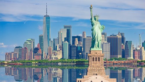 The Statue of Liberty stands in front of the New York City skyline, reflecting in calm waters under a blue sky.