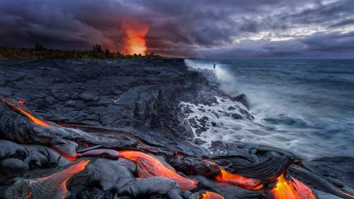 Lava flows into the ocean at dusk, with dramatic clouds and a distant eruption illuminating the sky.