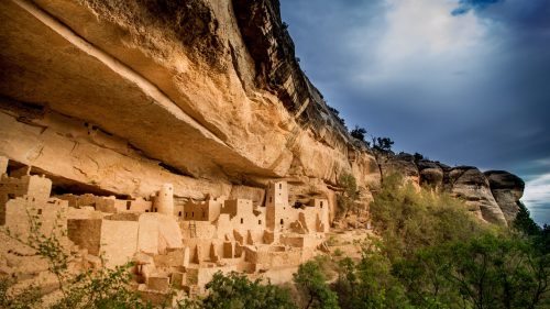 Ancient cliff dwellings nestled under a rocky overhang, surrounded by greenery and a dramatic sky.
