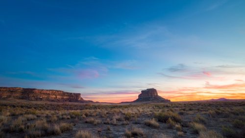 A vast desert landscape at sunset, featuring distant mesas and a colorful sky with soft clouds.