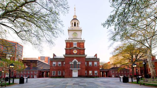 Historic building with a clock tower, surrounded by trees and a paved square, under a clear sky.