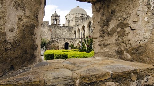 View through a stone archway showcasing a historic building with domes and lush greenery in the foreground.
