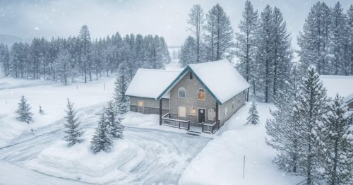 A cabin is in the middle of a snowy winter scene. Pine trees and mountains are covered in a blanket of snow as snowflakes fall to the ground.