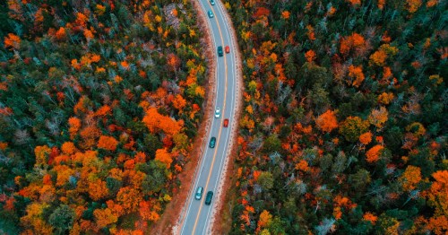 Aerial view of the winding Kancamagus Highway cutting through dense New Hampshire forest filled with vibrant autumn foliage in shades of orange, red, and gold.