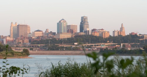 A city stands by a river with green grass in the foreground.