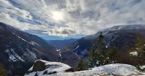 A vista of a mountain gorge covered in snow with the sun peeking through the clouds.