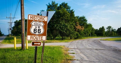 A brown Kansas Route 66 sign stands by the side of a road with green trees and grass.