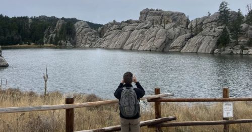A man is taking a photo of a lake with rock formations.