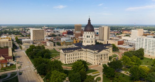 A domed building stands in the middle of a downtown with a blue sky and green trees.