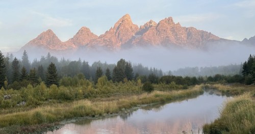 A layer of fog lies at the base of the Teton Mountains. A lazy river flows nearby.