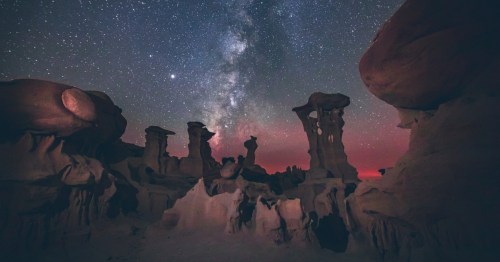 A starry night sky featuring the Milky Way arches over otherworldly hoodoo rock formations in a remote desert landscape, with a faint red glow on the horizon.