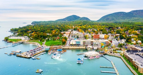 Aerial view of Bar Harbor, Maine, showing the waterfront harbor, marina, and colorful downtown buildings surrounded by fall foliage and mountains near Acadia National Park.