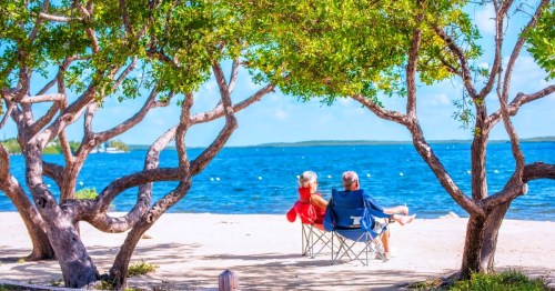Two vacationers sitting in folding chairs on the sand shores of a beach in Florida, surrounded by two trees curved with branches.