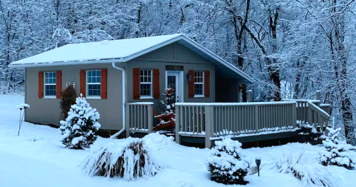 An exterior view of a cozy Airbnb cottage in Rushford, MN, surrounded by a forest heavily coated in fresh white snow during a serene winter evening.