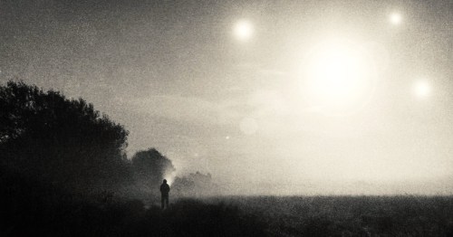 A man standing in a field holding a flashlight staring at mysterious bright lights in the sky on a hazy night.