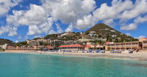 The beachfront at Divi Carina Bay Resort features expansive white sand lined with blue lounge chairs and umbrellas, overlooking turquoise waters with resort buildings and a lush green hill in the background.