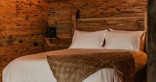 Wood walled cabin bedroom with bed with white sheets and a brown, fuzzy blanket