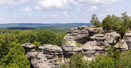 Rock formations tower over a green forest with a blue sky.